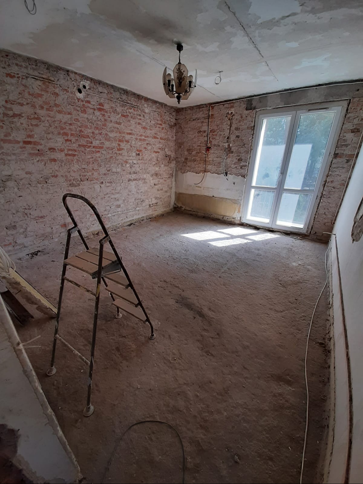 Pokój w trakcie remontu: odsłonięta cegła, stara instalacja elektryczna, drabina, okno z widokiem na zieleń. Przygotowanie do dalszych prac wykończeniowych.