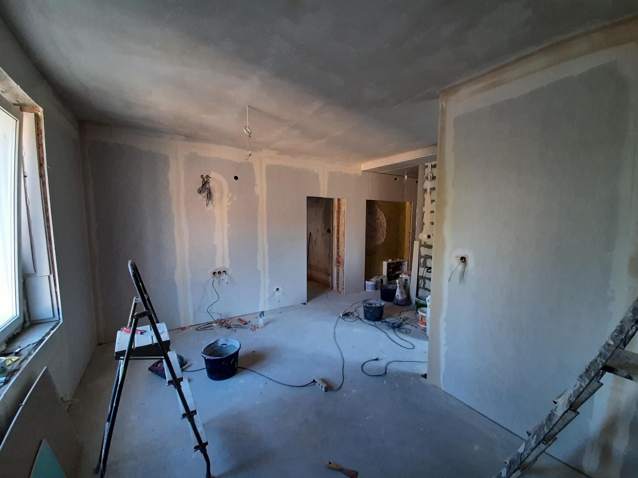 Wnętrze w trakcie prac wykończeniowych: ściany z płyt karton-gips, częściowo zagruntowane, widoczne kable, drabina i narzędzia malarskie. Przygotowanie do dalszych prac.