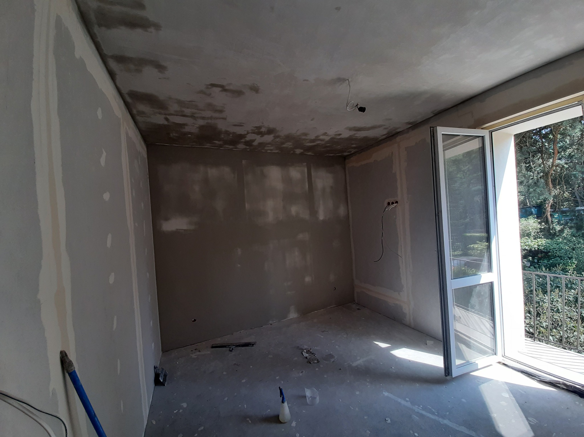 Pokój w trakcie prac wykończeniowych: ściany z płyt karton-gips, surowy sufit, otwarte drzwi balkonowe wpuszczające światło. Widoczne narzędzia na podłodze.