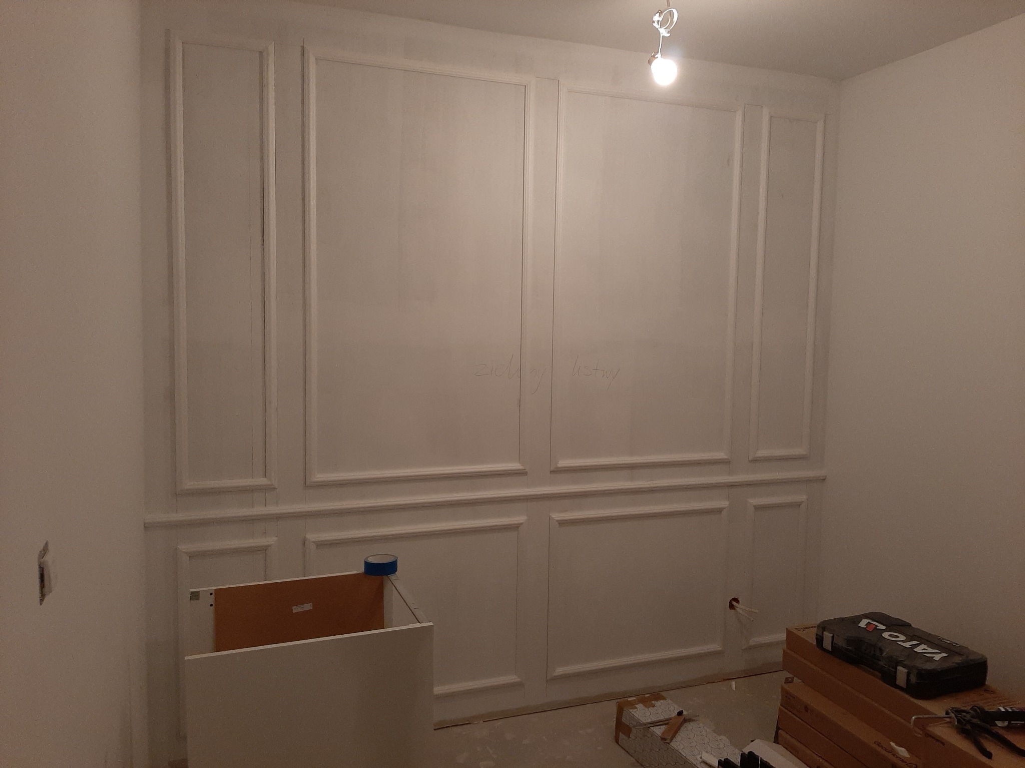 Biała ściana z dekoracyjnymi listwami w stylu klasycznym, częściowo zamontowanymi. Widoczne narzędzia i materiały budowlane, sugerujące etap prac wykończeniowych.