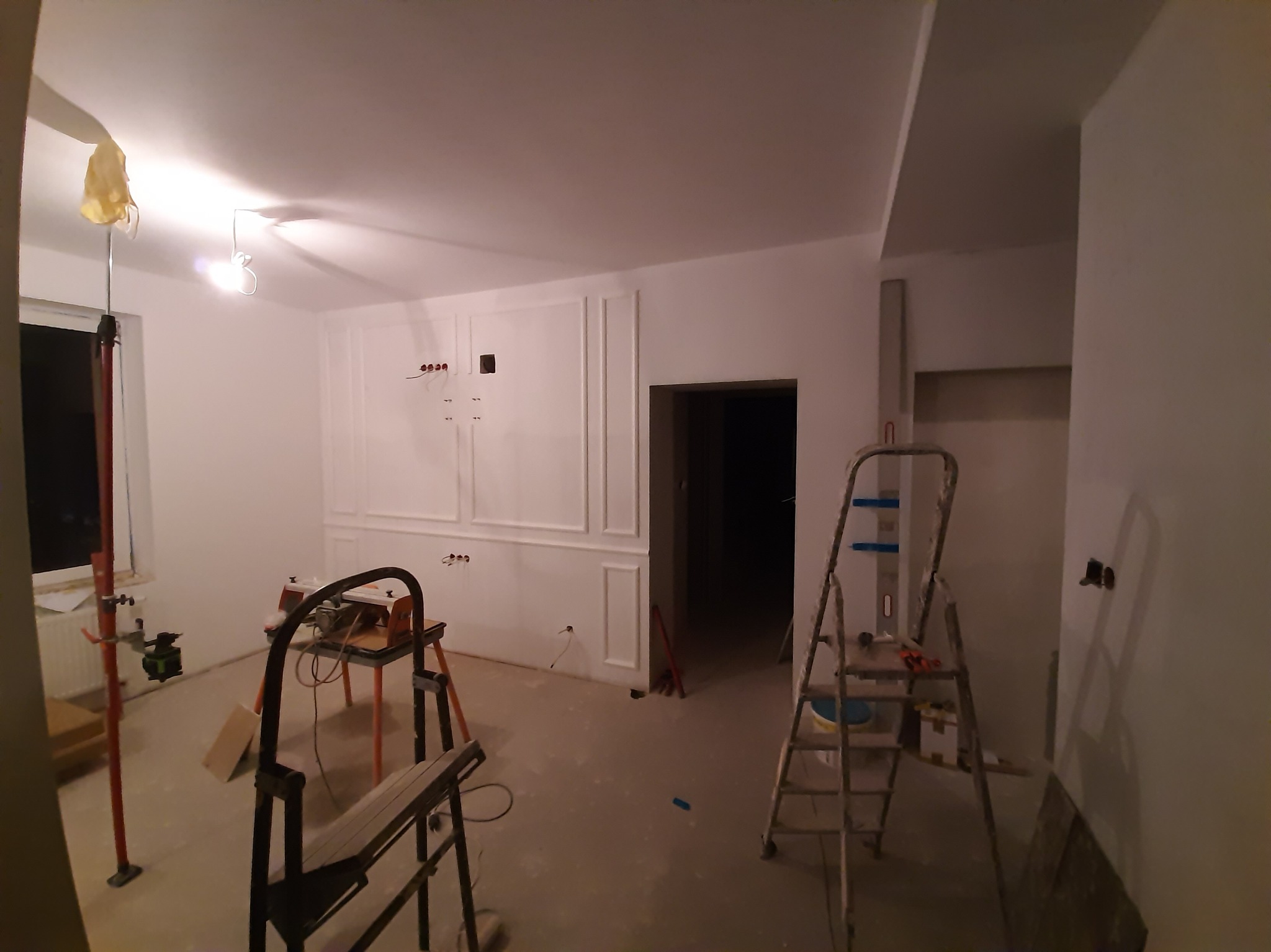 Białe ściany z dekoracyjnymi panelami w trakcie montażu, widoczne narzędzia i drabina, surowy stan podłogi, przygotowania do wykończenia wnętrza.