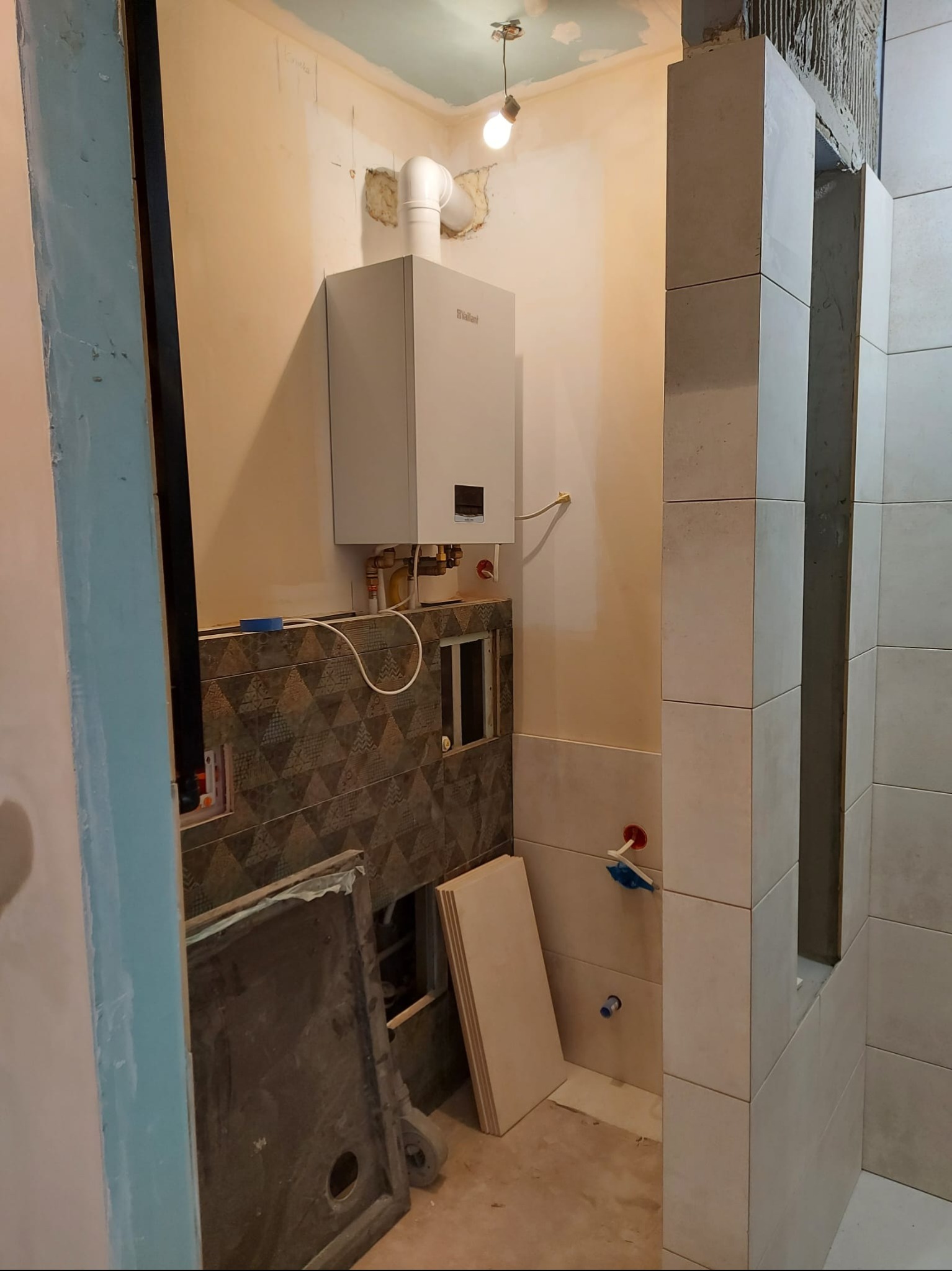 Remont łazienki w trakcie: częściowo ułożone płytki na ścianie, zamontowany piec gazowy, widoczne materiały budowlane i instalacje. Wnętrze w stanie surowym.