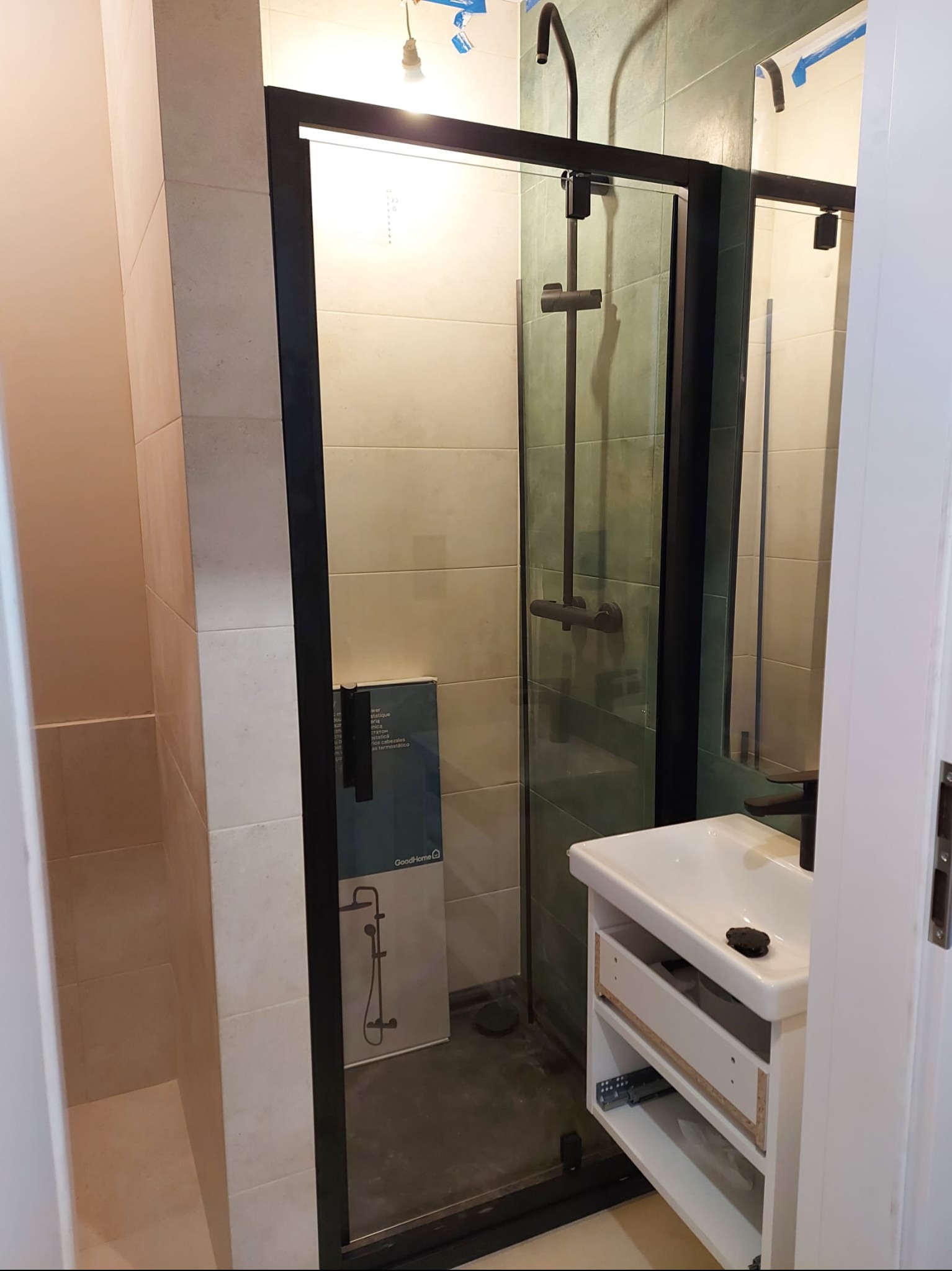 Nowoczesna kabina prysznicowa z czarną ramą i deszczownicą, umywalka z szafką w trakcie montażu, jasne płytki na ścianach. Widoczny karton z grafiką baterii.