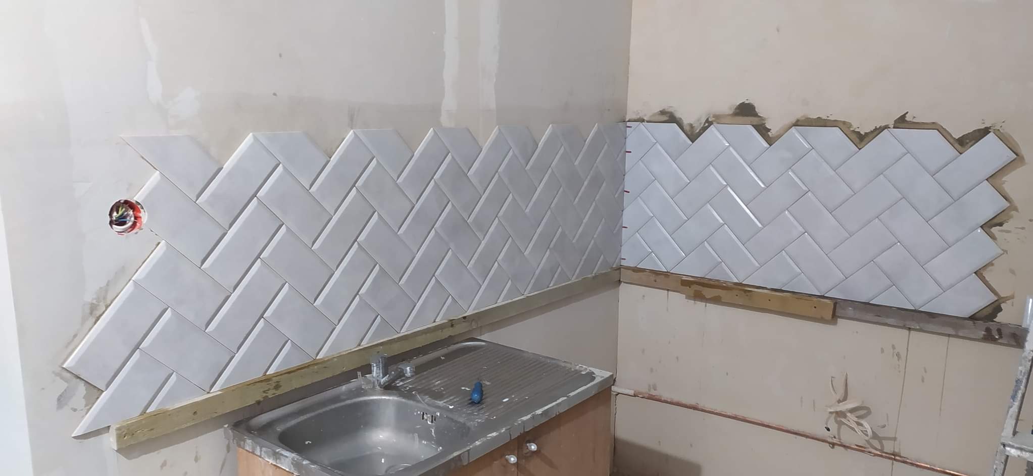 Układanie białych, błyszczących płytek w jodełkę na ścianie nad zlewem w trakcie prac. Widoczne listwy pomocnicze i niedokończone krawędzie.