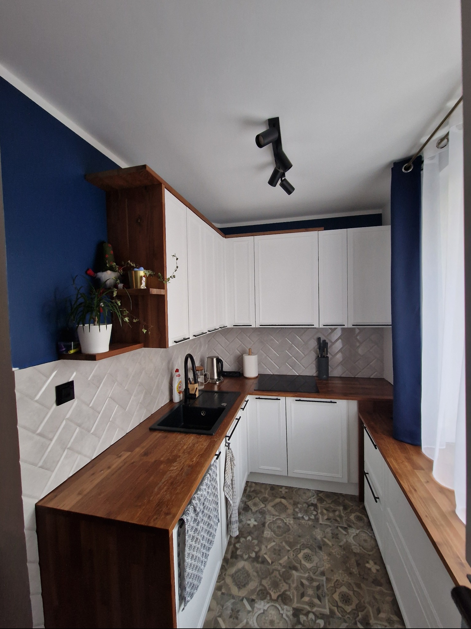 Nowoczesna kuchnia w odcieniach bieli, drewna i granatu. Uwagę przykuwają drewniane blaty, białe szafki z frezowanymi frontami i wzorzysta podłoga.