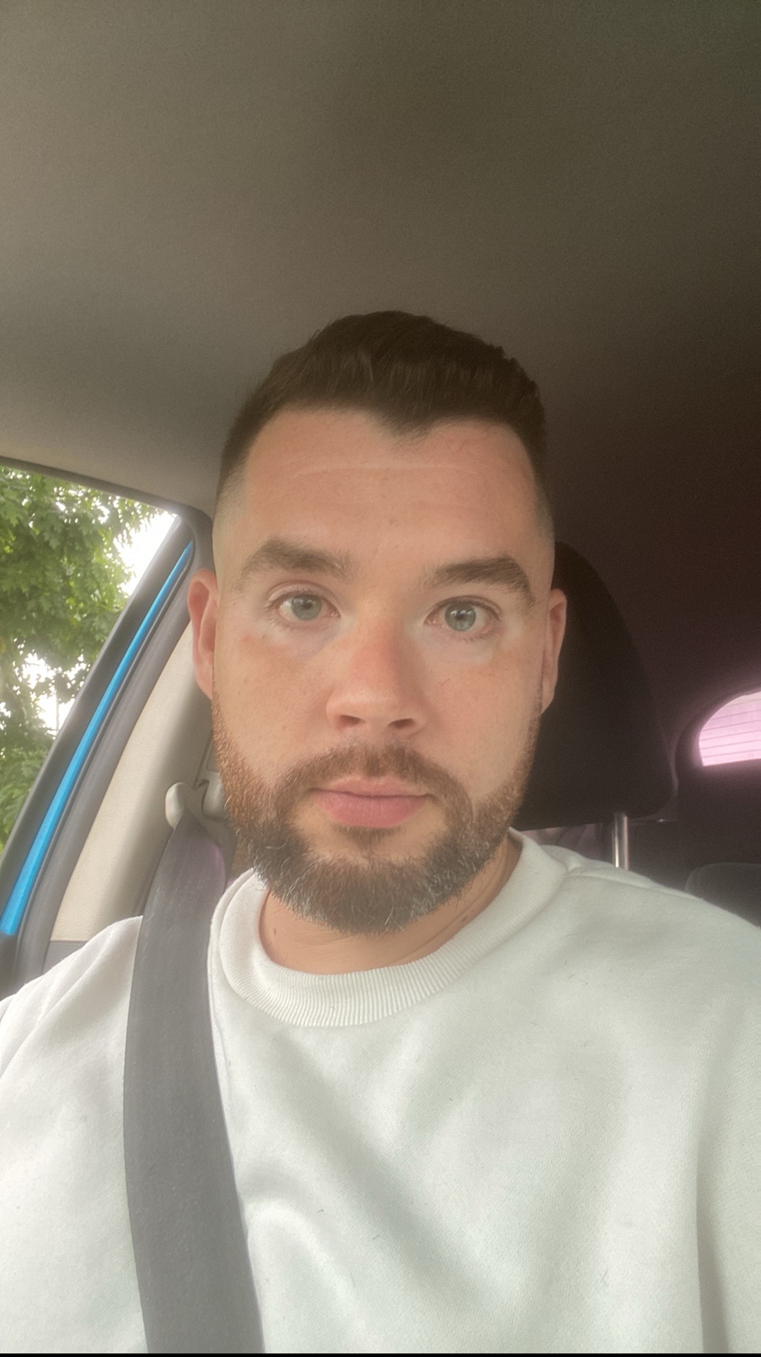 Mężczyzna z brodą w białej bluzie, siedzący w samochodzie z zapiętym pasem. Widok z perspektywy kierowcy, światło dzienne.