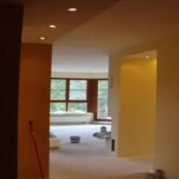 Wnętrze w trakcie remontu: świeżo pomalowane ściany w odcieniach beżu i żółci, widoczne puszki z farbą, surowe wykończenie podłogi, okno z widokiem na zieleń.