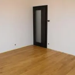 Pusty pokój z parkietem w odcieniach miodu i białymi ścianami. W rogu ciemne drzwi z matową szybą i czarną klamką. Widoczne dwa otwory w ścianie po prawej stronie.