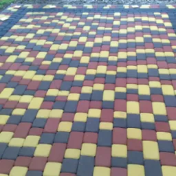Ułożona nawierzchnia z kostki brukowej w odcieniach żółci, czerwieni i grafitu, ułożona we wzór szachownicy, widoczne białe znaczniki na obrzeżach.