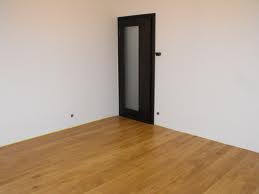 Pusty pokój z parkietem w odcieniach miodu i białymi ścianami. W rogu ciemne drzwi z matową szybą i czarną klamką. Widoczne dwa otwory w ścianie po prawej stronie.