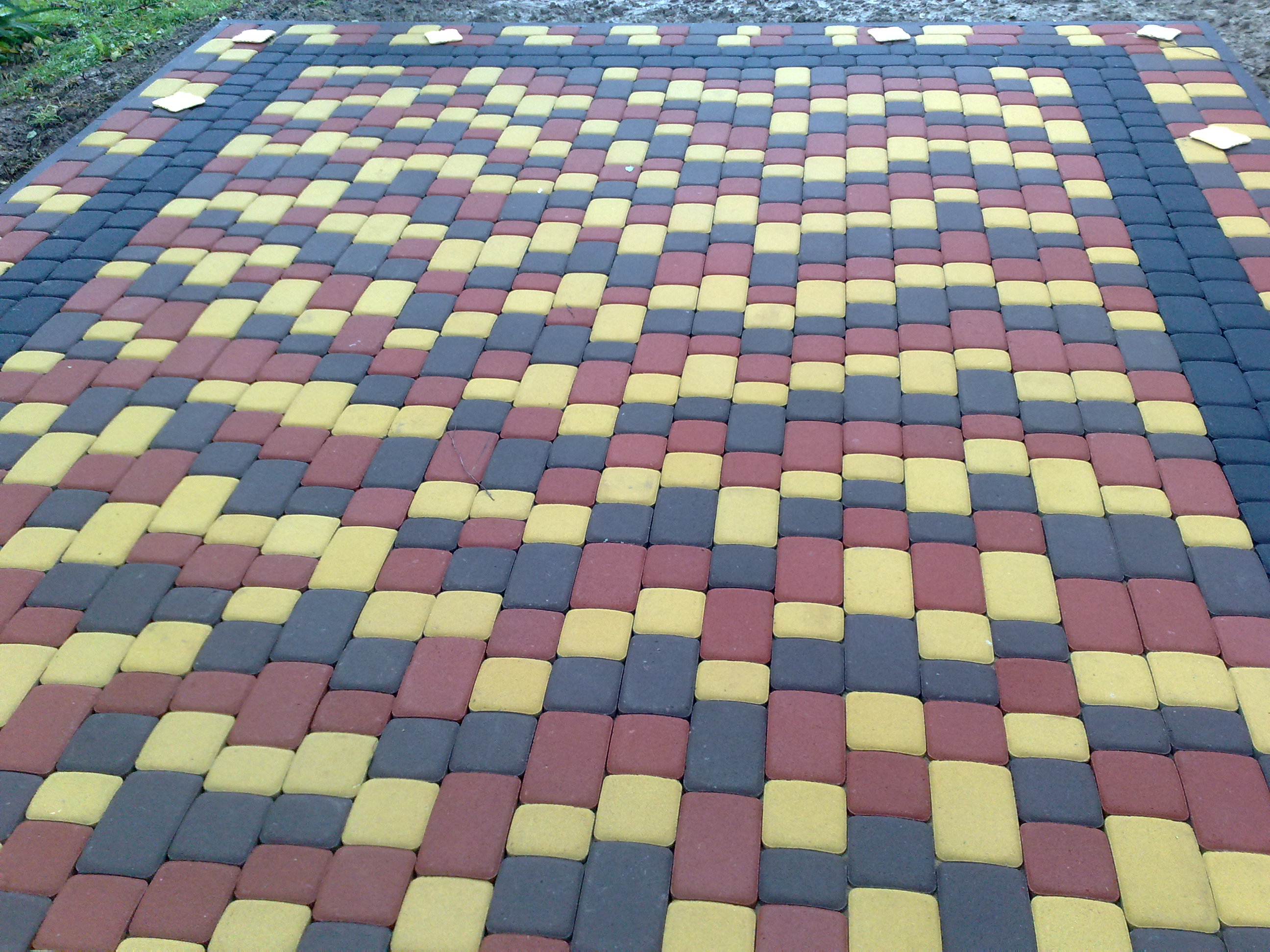 Ułożona nawierzchnia z kostki brukowej w odcieniach żółci, czerwieni i grafitu, ułożona we wzór szachownicy, widoczne białe znaczniki na obrzeżach.