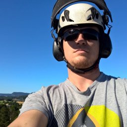 Tehtree - Autoportret pracownika w kasku i okularach ochronnych na tle błękitnego nieba i zielonego krajobrazu. Widoczne drobne trociny na koszulce.