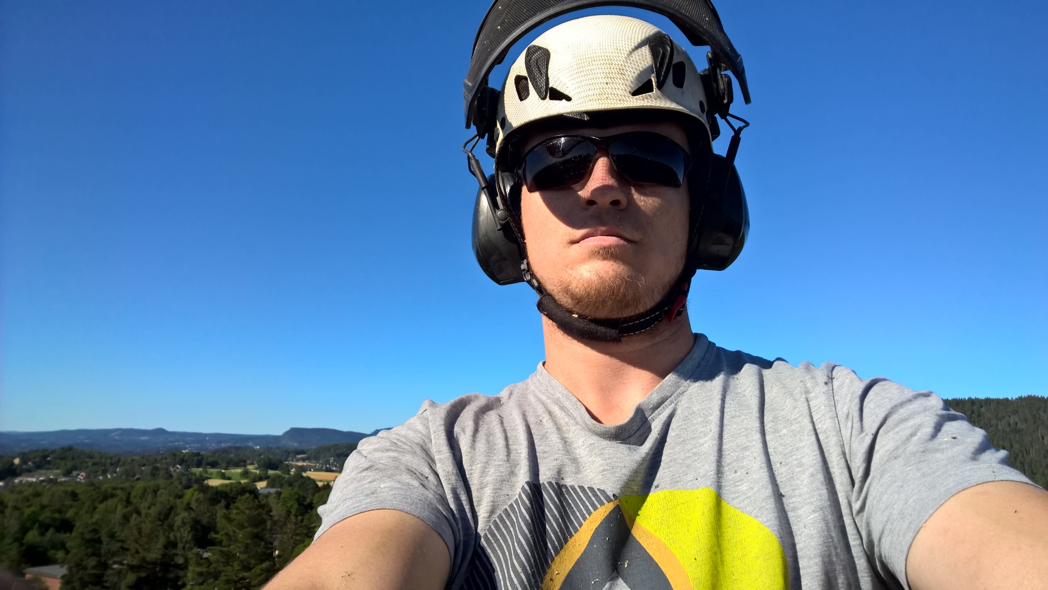 Autoportret pracownika w kasku i okularach ochronnych na tle błękitnego nieba i zielonego krajobrazu. Widoczne drobne trociny na koszulce.