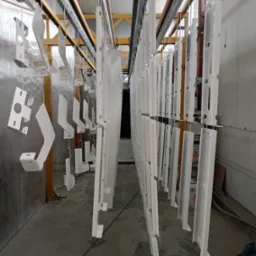 Białe metalowe elementy zawieszone na linkach w hali, prawdopodobnie po procesie malowania, z widoczną konstrukcją sufitową i oknami w tle.