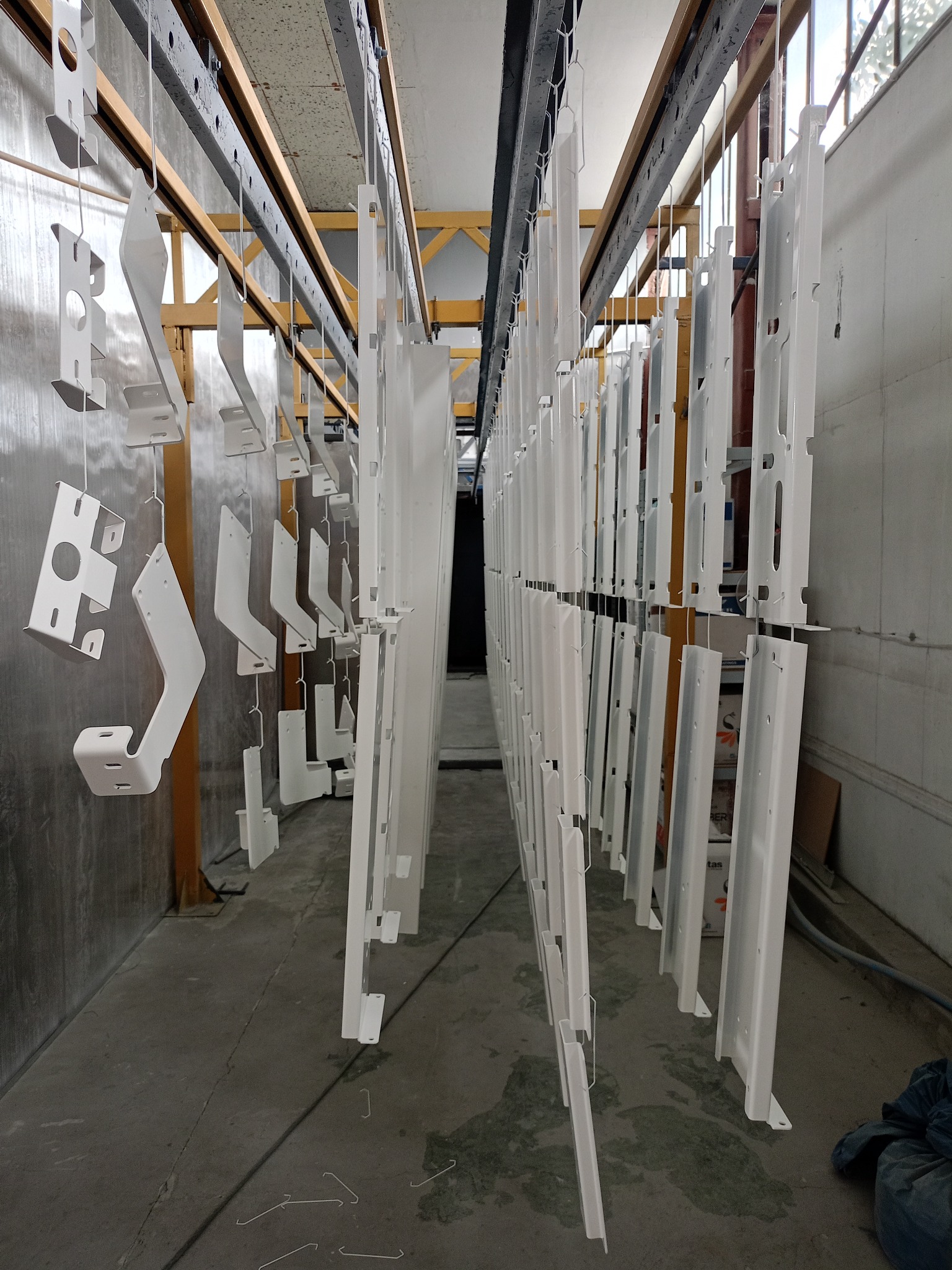Białe metalowe elementy zawieszone na linkach w hali, prawdopodobnie po procesie malowania, z widoczną konstrukcją sufitową i oknami w tle.