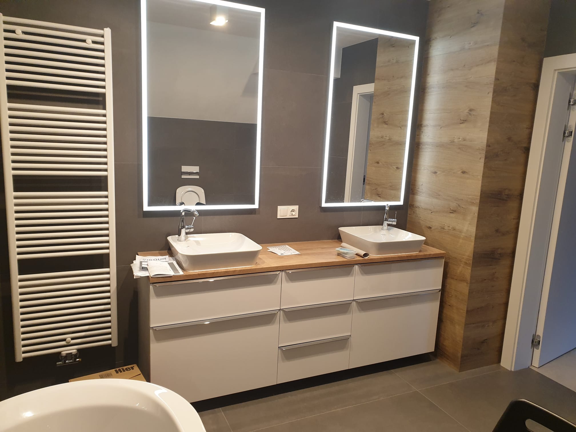 Nowoczesna łazienka z dwoma umywalkami nablatowymi, podświetlanymi lustrami i grzejnikiem drabinkowym. Umywalki na drewnianym blacie i szafkach z szufladami.