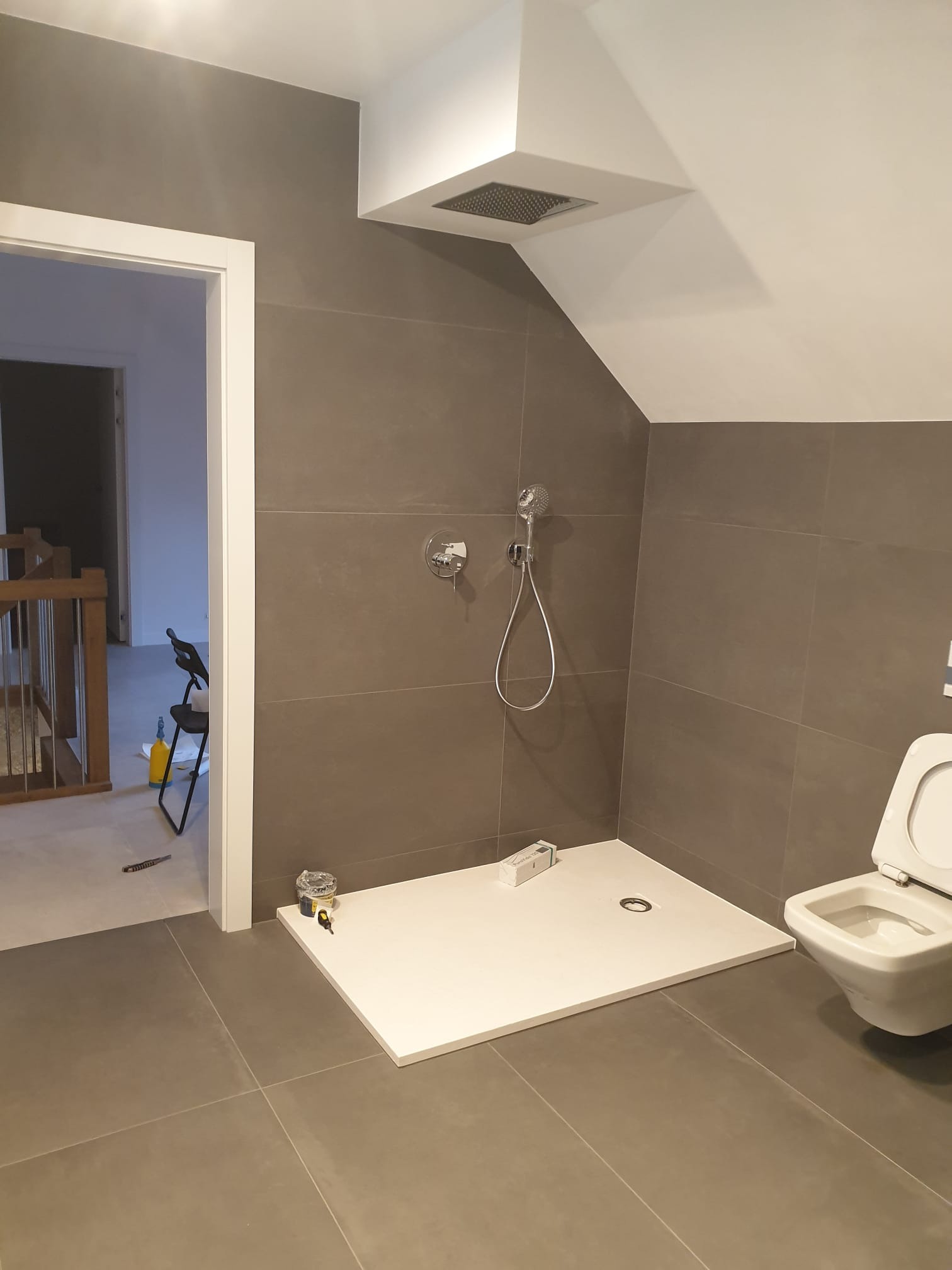 Nowoczesna łazienka z szarymi płytkami, białą kabiną prysznicową i toaletą. Widoczny fragment korytarza z drewnianymi schodami. Oświetlenie sufitowe.