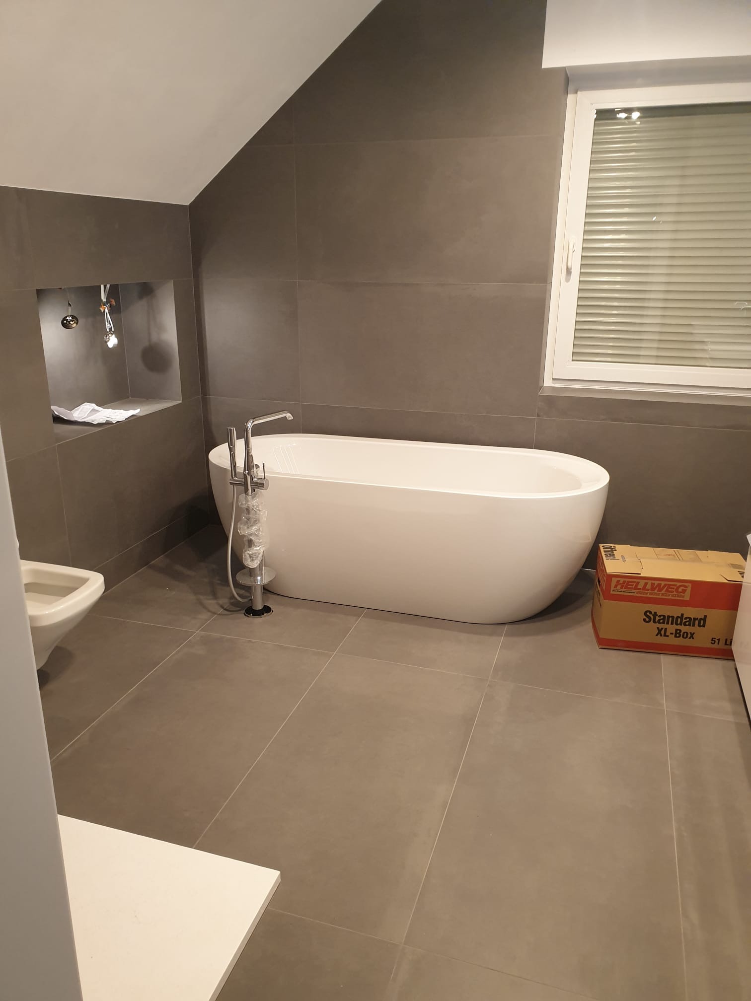 Nowoczesna łazienka z wolnostojącą wanną na tle szarych płytek, z oknem z żaluzjami i przygotowanym miejscem na umywalkę. Widoczna kartonowa skrzynka.