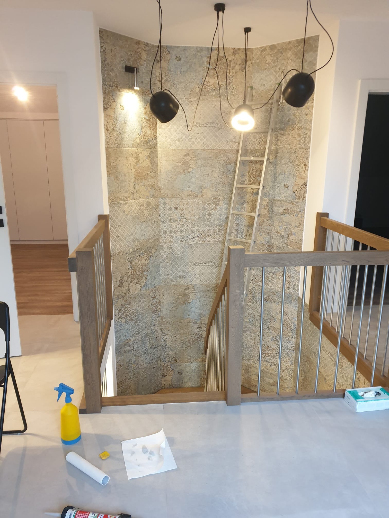 Wykończenie klatki schodowej z dekoracyjną ścianą z płytek i drewnianą balustradą. Widoczne narzędzia i drabina sugerują trwający proces wykończeniowy.