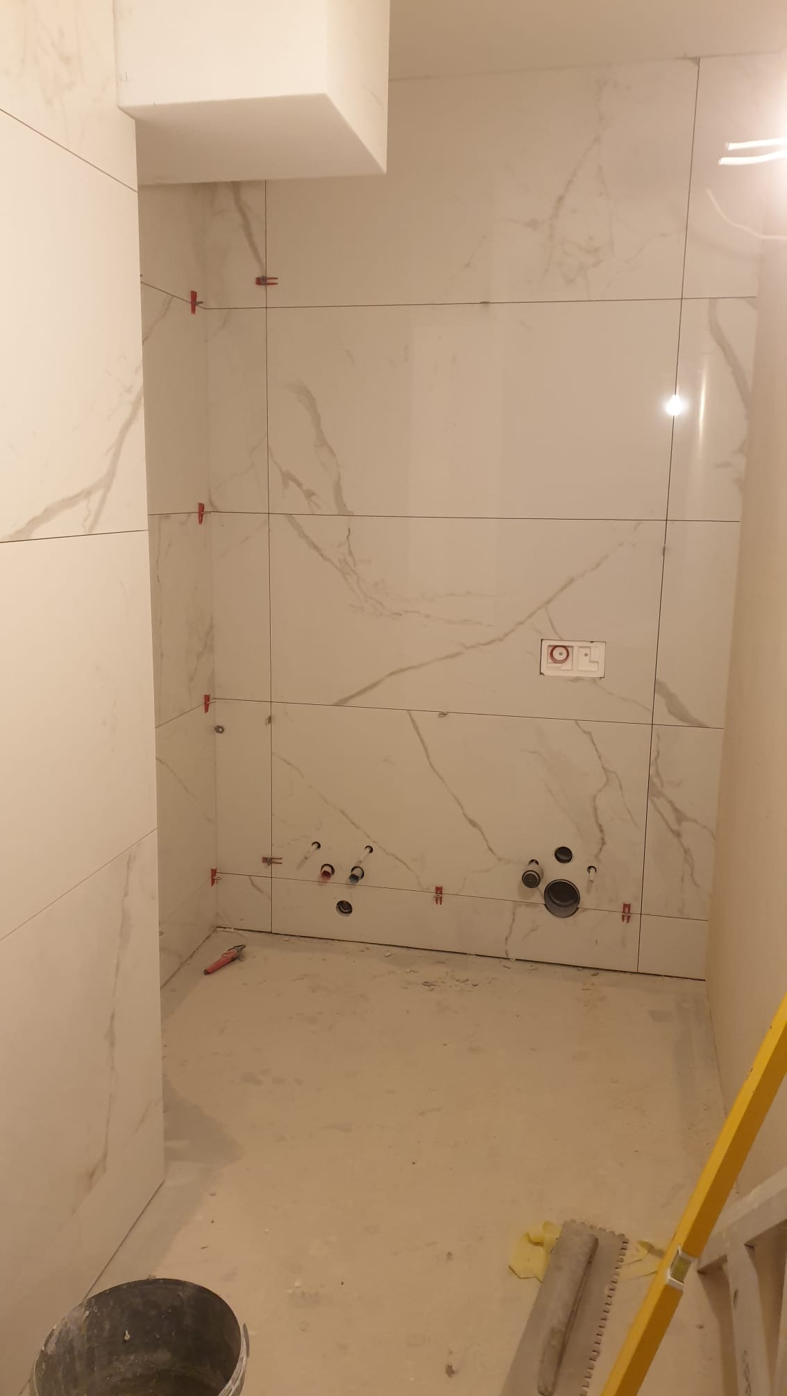 Białe płytki imitujące marmur na ścianach łazienki w trakcie montażu. Widoczne krzyżyki dystansowe, otwory na instalacje i wiadro. Żółta poziomica w rogu.