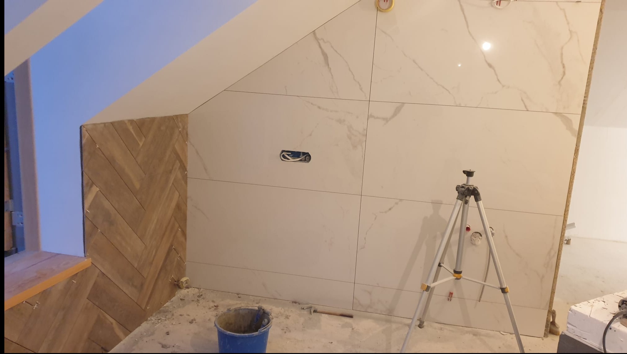 Układanie płytek imitujących marmur na ścianie pod skosem, obok fragmentu ściany wykończonej drewnopodobną mozaiką w jodełkę. Widoczny sprzęt glazurniczy.