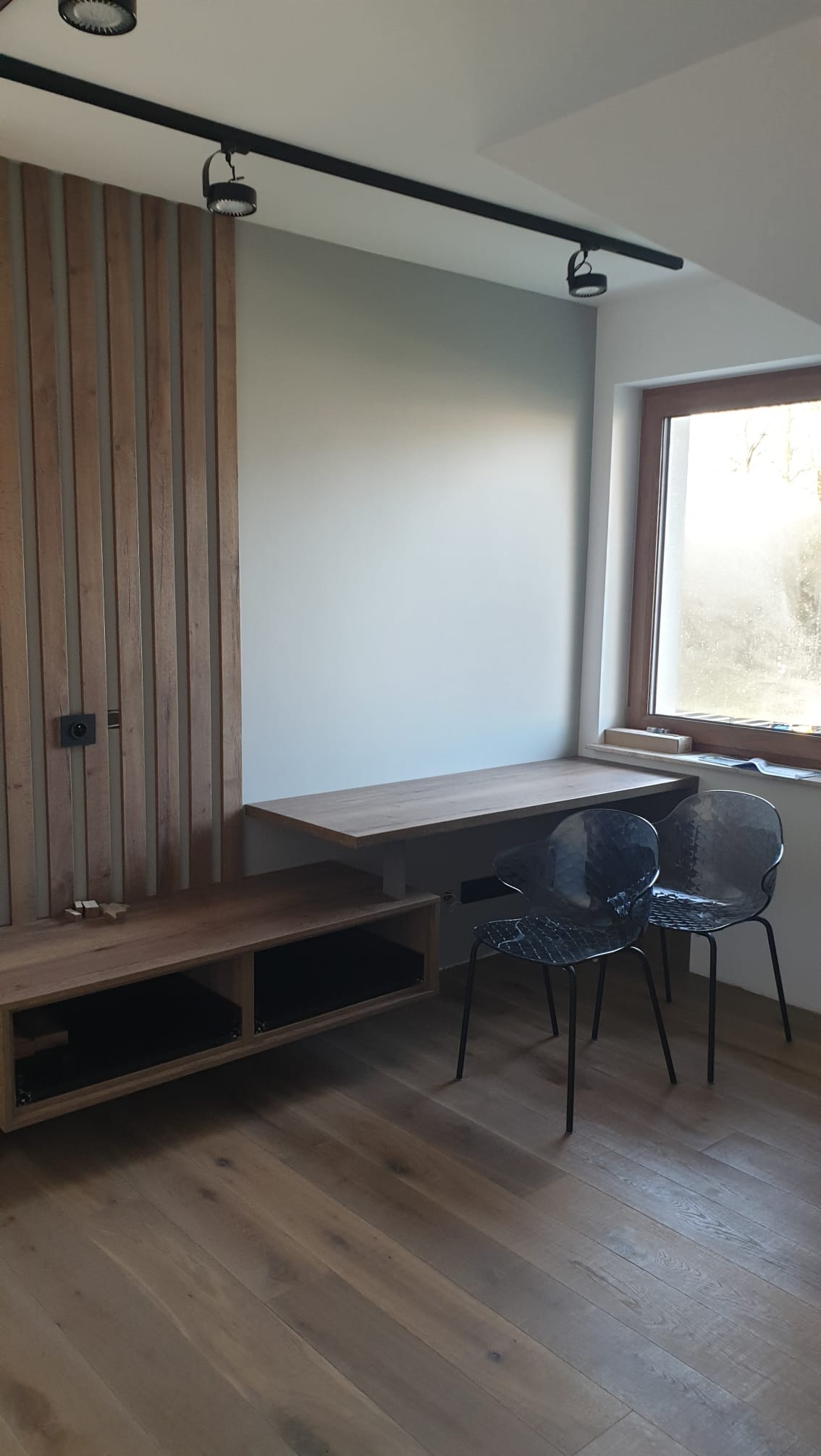 Nowoczesne wykończenie wnętrza: drewniana zabudowa ściany z półkami, biurko pod oknem i podłoga z paneli. Dwa krzesła w tle. Oświetlenie szynowe.