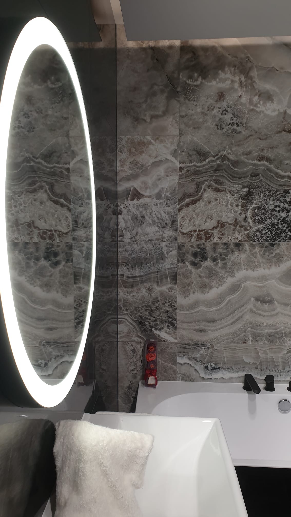 Wykończona łazienka z owalnym lustrem z podświetleniem LED, marmurowymi płytkami i białą wanną z czarną armaturą. Dekoracyjny akcent: pudełko z różami.