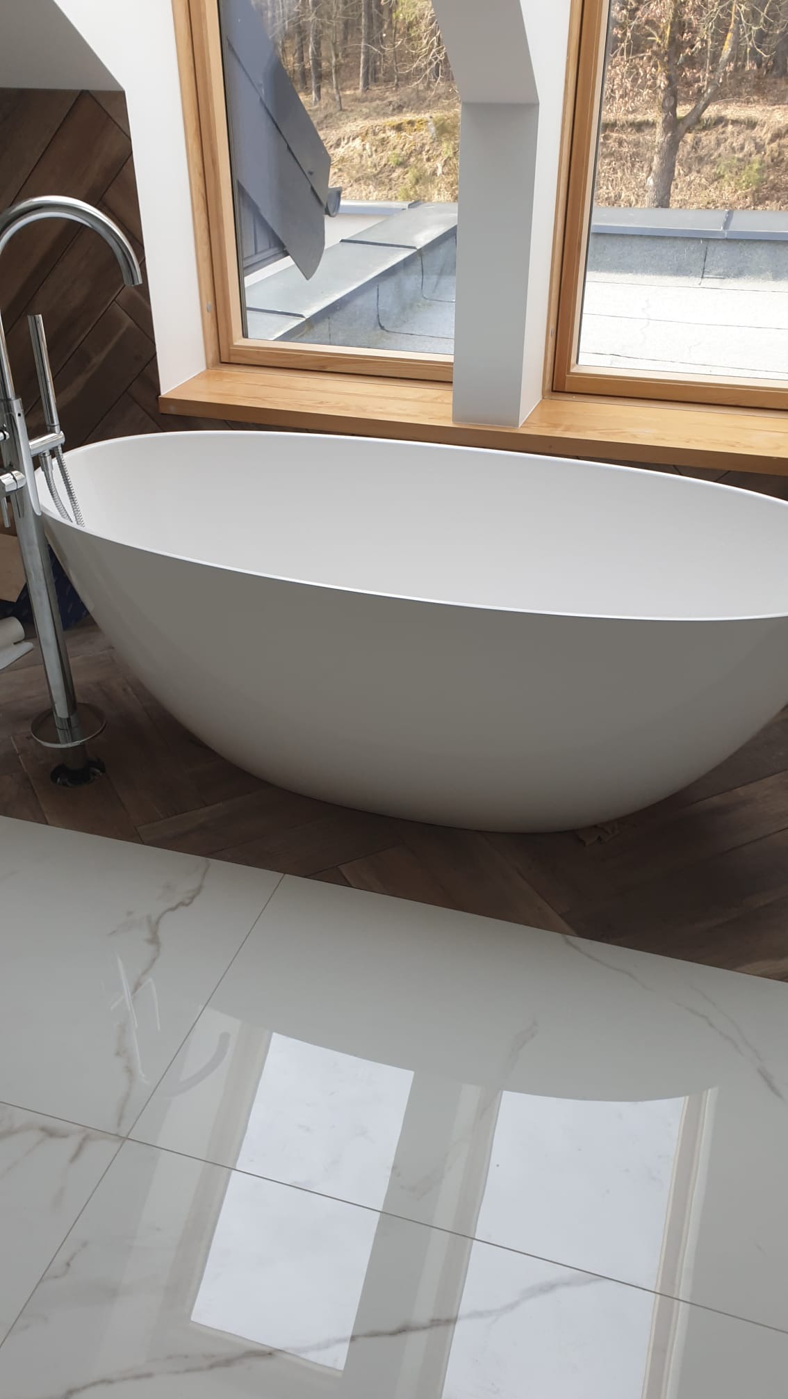 Wolnostojąca wanna owalna z chromowaną armaturą na tle okna połaciowego i parkietu ułożonego w jodełkę. Nowoczesna łazienka z marmurową posadzką.