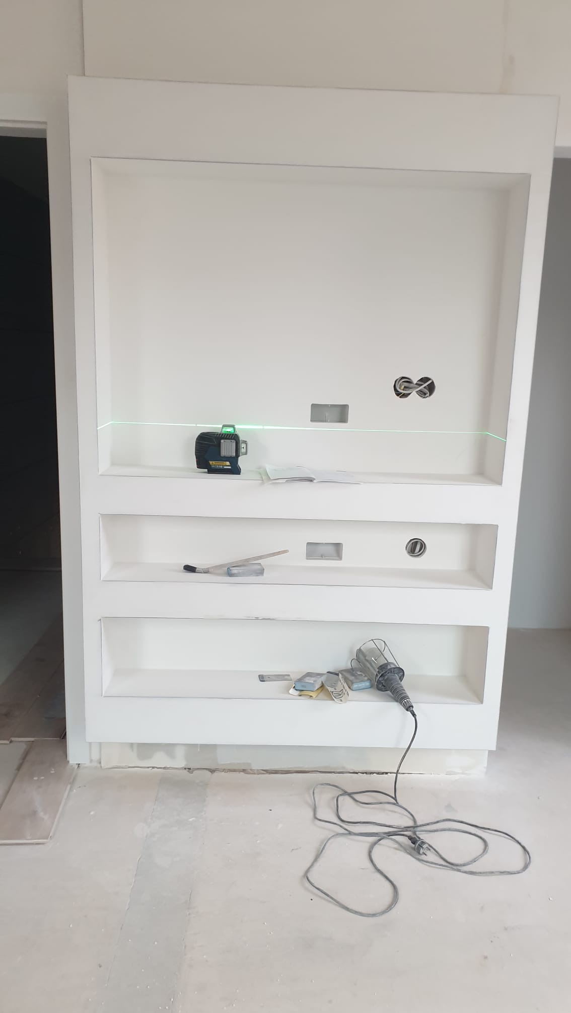 Biała, wnękowa półka z trzema poziomami, oświetlona laserem krzyżowym. Widoczne narzędzia i gniazdka elektryczne. Detal wykończenia wnętrza w trakcie prac.