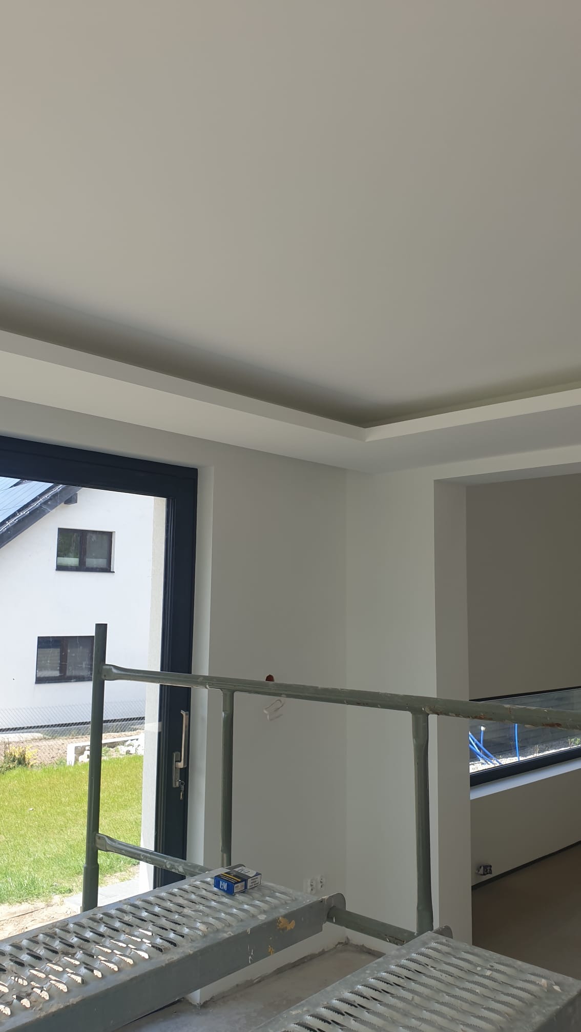 Wnętrze po malowaniu: białe ściany, sufit z dekoracyjnym obramowaniem, fragment okna z widokiem na dom i trawnik, rusztowanie malarskie w pomieszczeniu.