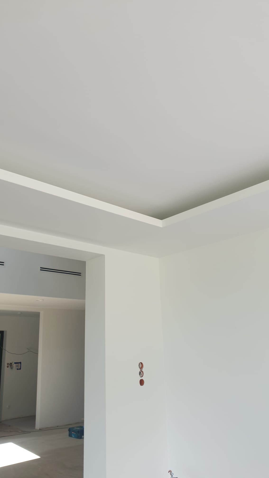 Minimalistyczne wykończenie wnętrza: białe ściany, sufit podwieszany z oświetleniem, widoczne gniazdka elektryczne, częściowo widoczne podłoże i przejście do innego pomieszczenia.