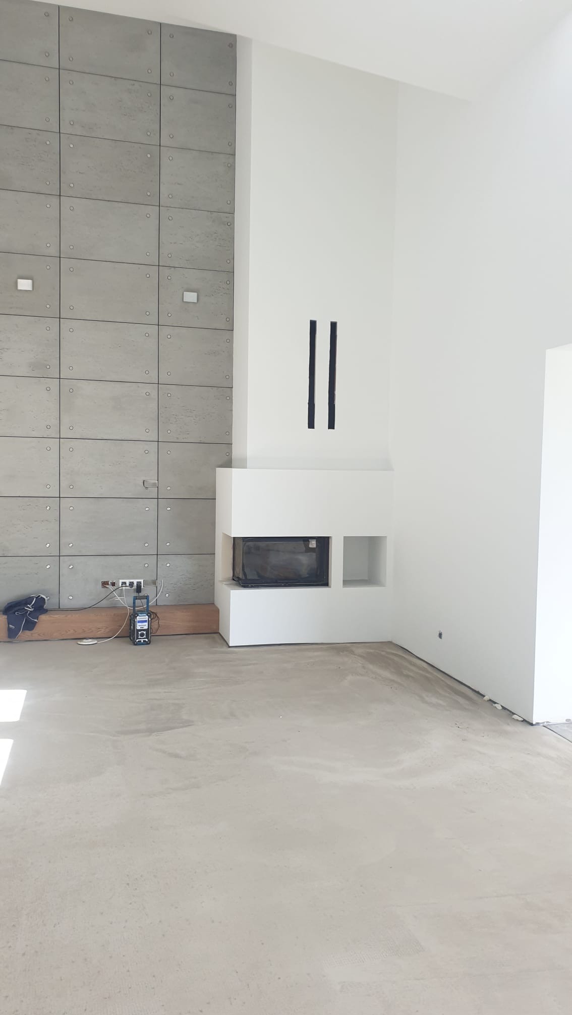 Surowe wnętrze z betonową ścianą, kominkiem i gładką posadzką. Widoczne narzędzia i elementy instalacji elektrycznej. Minimalistyczny design w szarościach i bieli.