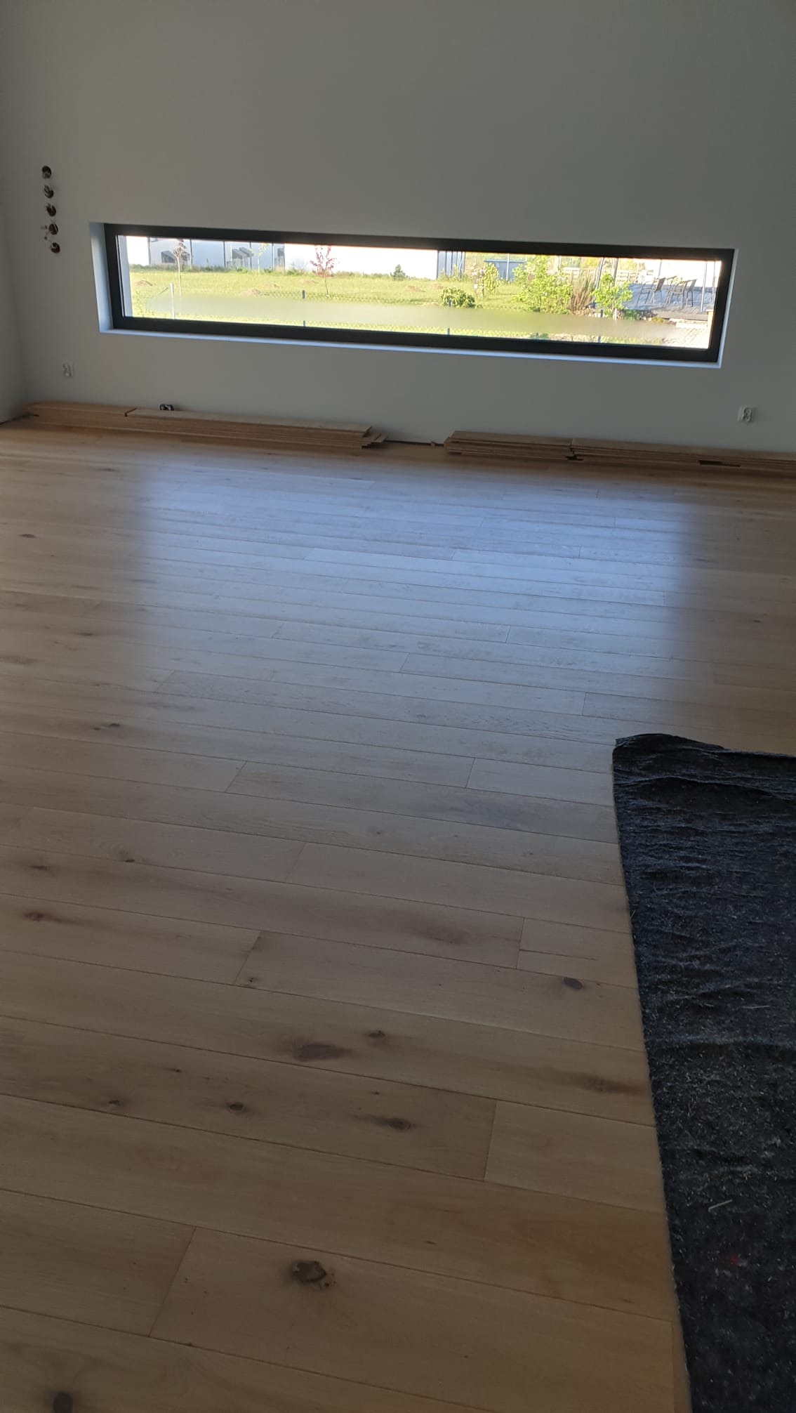 Jasny, drewniany parkiet ułożony w nowym wnętrzu z poziomym oknem. Fragment czarnej maty ochronnej w rogu. W tle widok na zieleń.