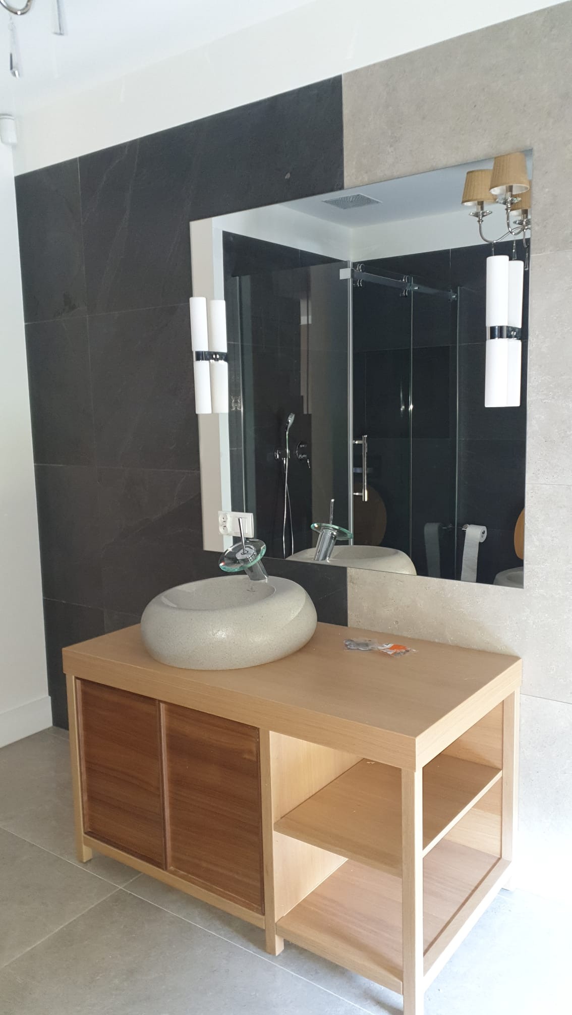 Nowoczesna łazienka z umywalką w kształcie kamienia na drewnianej szafce. W tle lustro odbijające kabinę prysznicową i czarne płytki na ścianie.