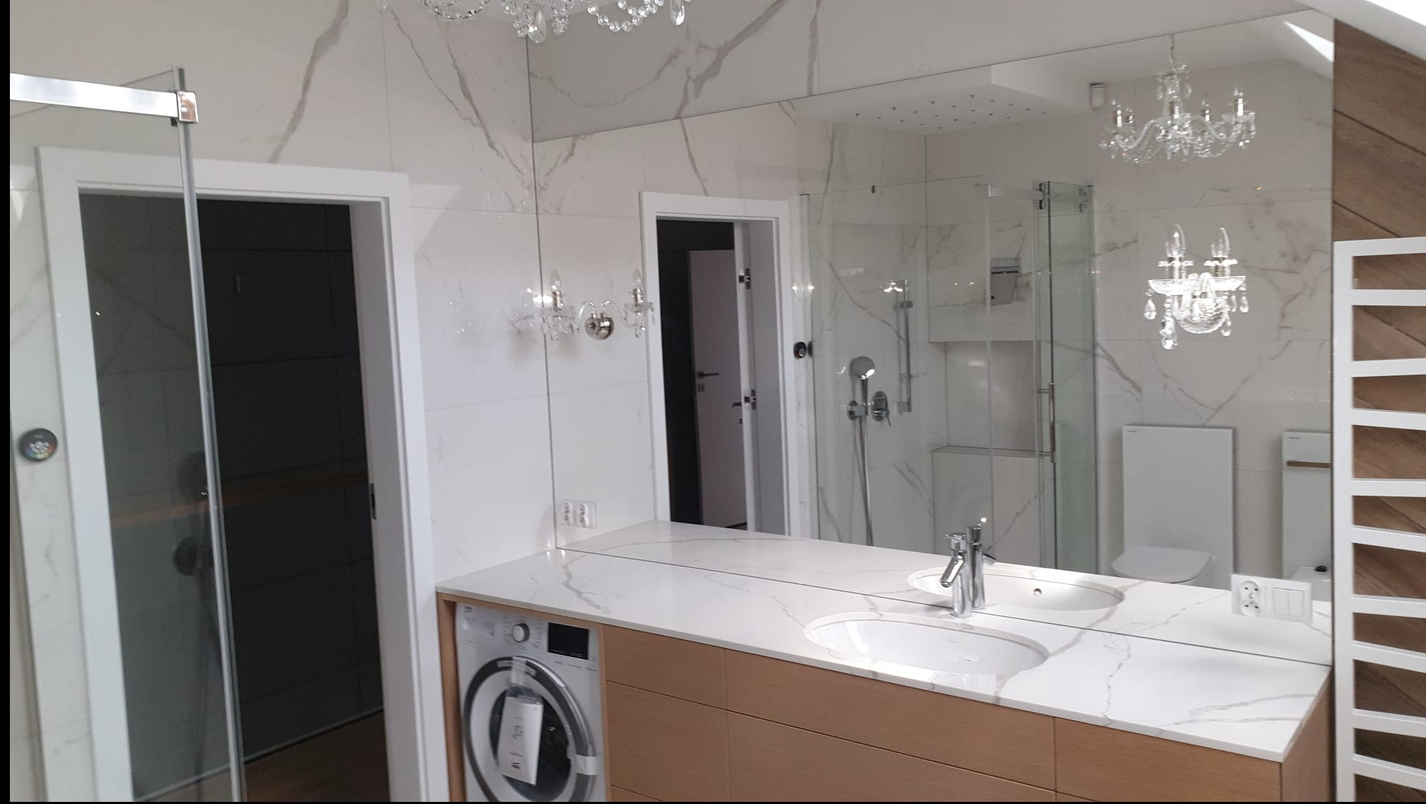 Elegancka łazienka z marmurowymi płytkami, dużym lustrem, drewnianą szafką pod umywalką i pralką w zabudowie. Widoczny prysznic za szklaną ścianą.