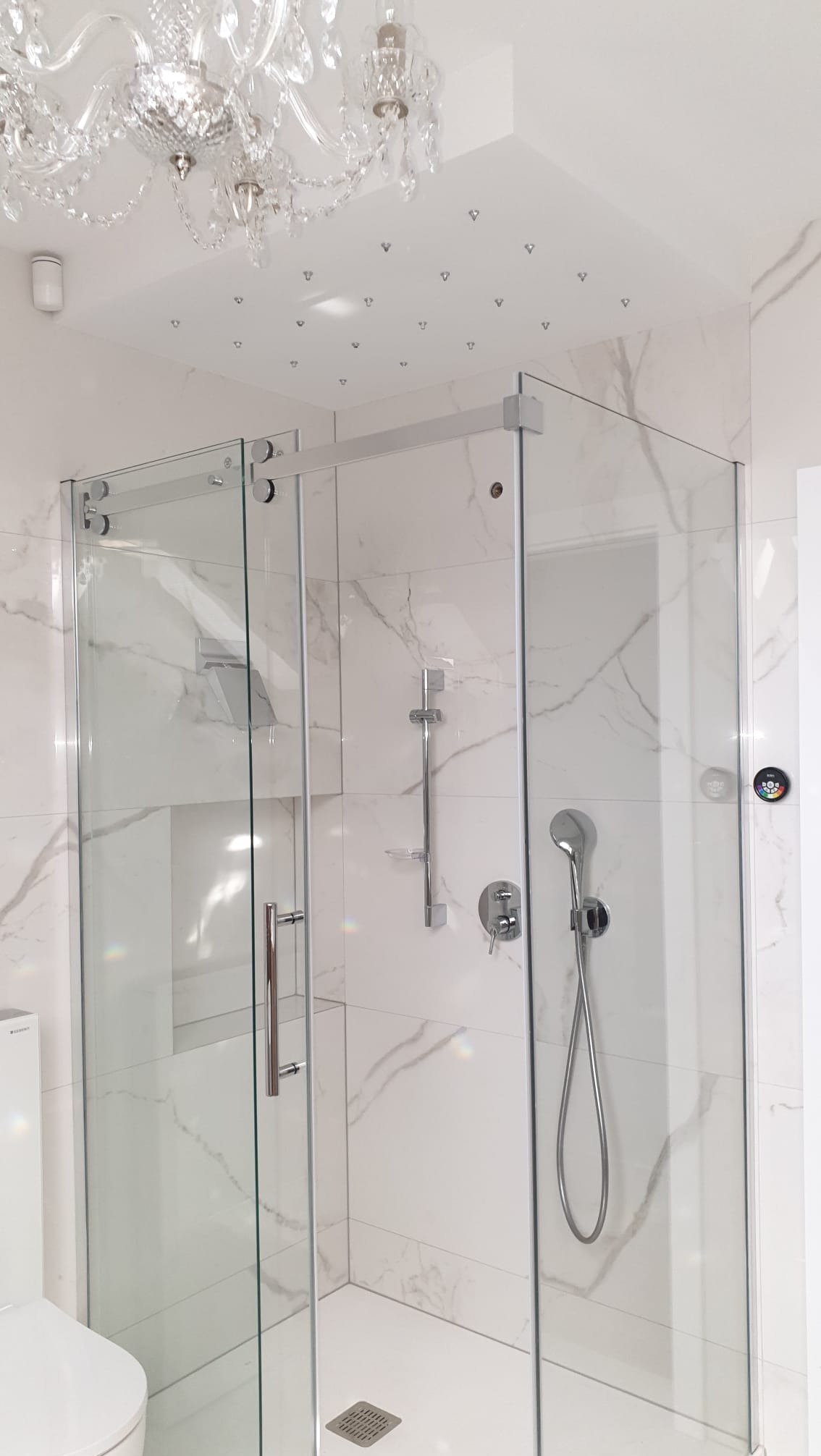 Nowoczesna kabina prysznicowa z marmurowymi płytkami i kryształowym żyrandolem. Chromowane detale, minimalistyczny design, eleganckie wykończenie łazienki.