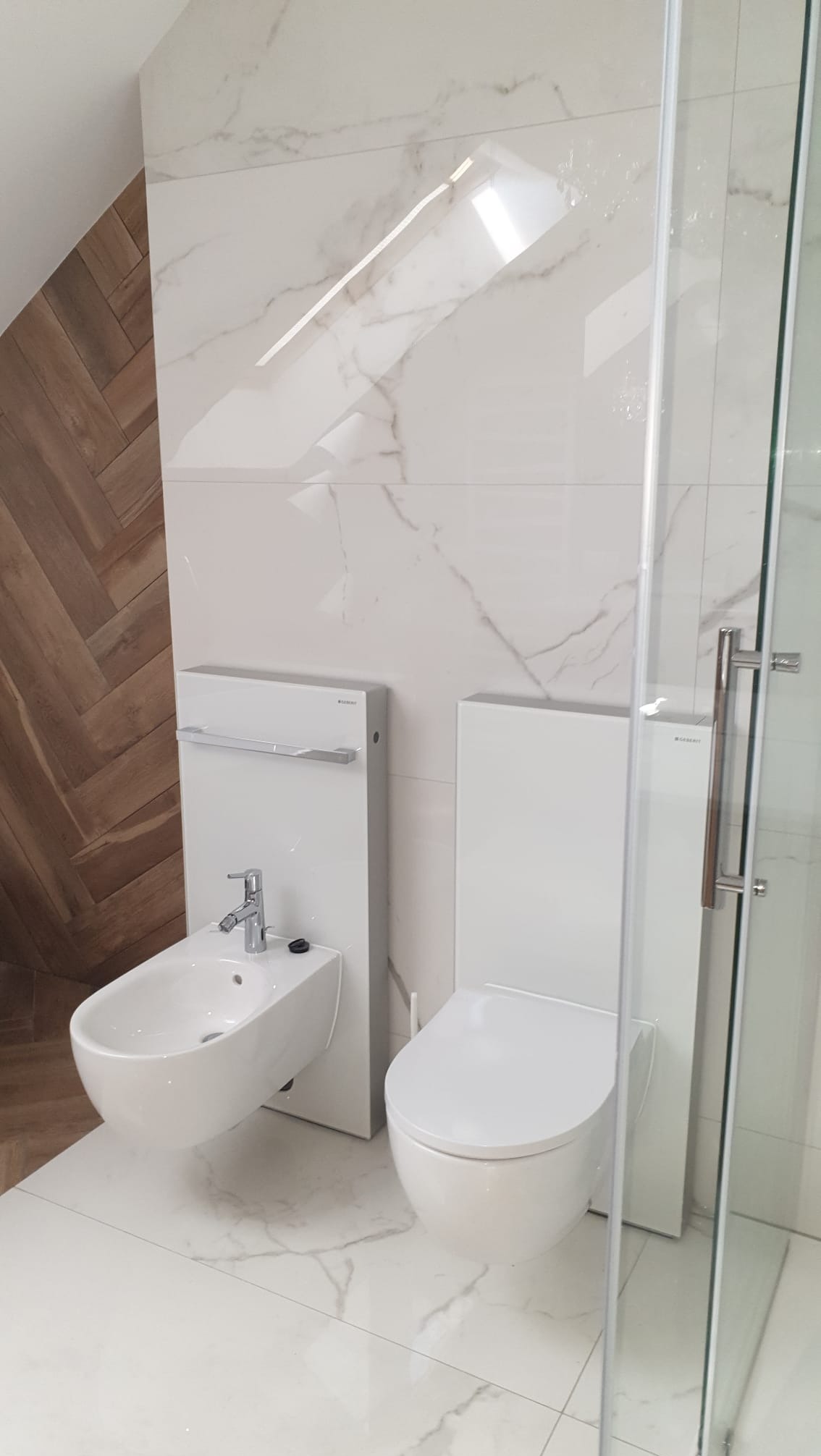 Nowoczesna łazienka z białymi płytkami imitującymi marmur, wiszącą toaletą i bidetem. Na ścianie drewniana okładzina w jodełkę. Szklana kabina prysznicowa z chromowanym uchwytem.