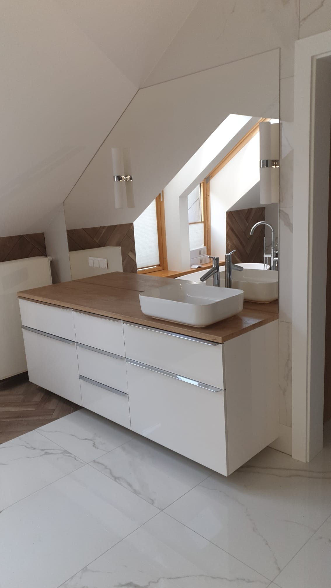 Nowoczesna łazienka na poddaszu: białe szafki z drewnianym blatem, prostokątna umywalka, duże lustro, marmurowe płytki podłogowe i okno dachowe.