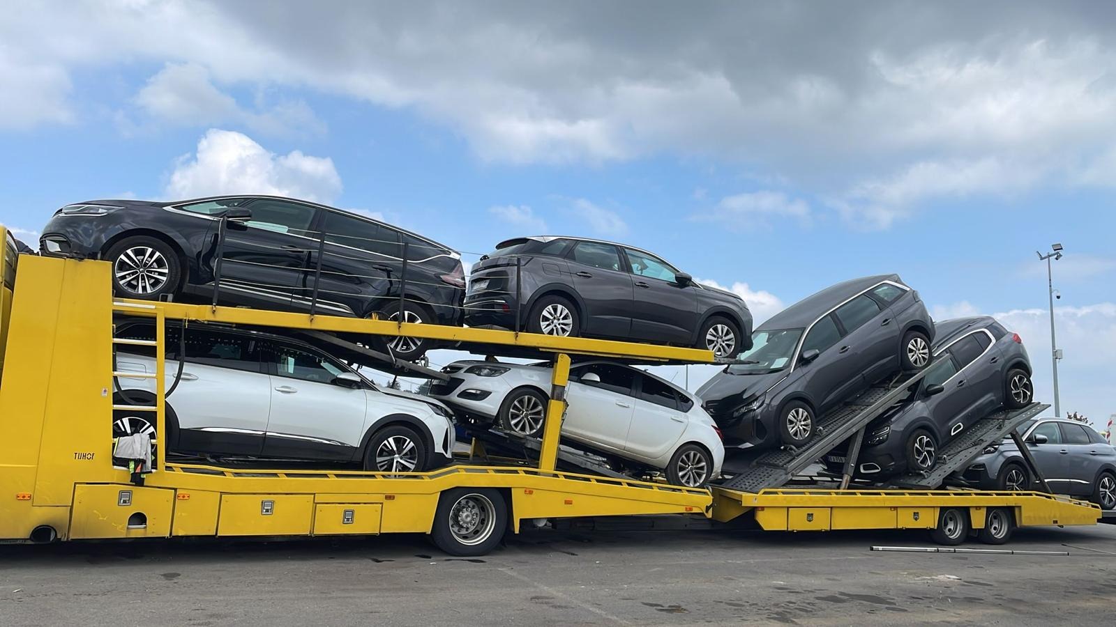 Żółta laweta z załadowanymi samochodami osobowymi różnych marek i kolorów na tle pochmurnego nieba. Widok z dołu, perspektywa lekko zniekształcona.