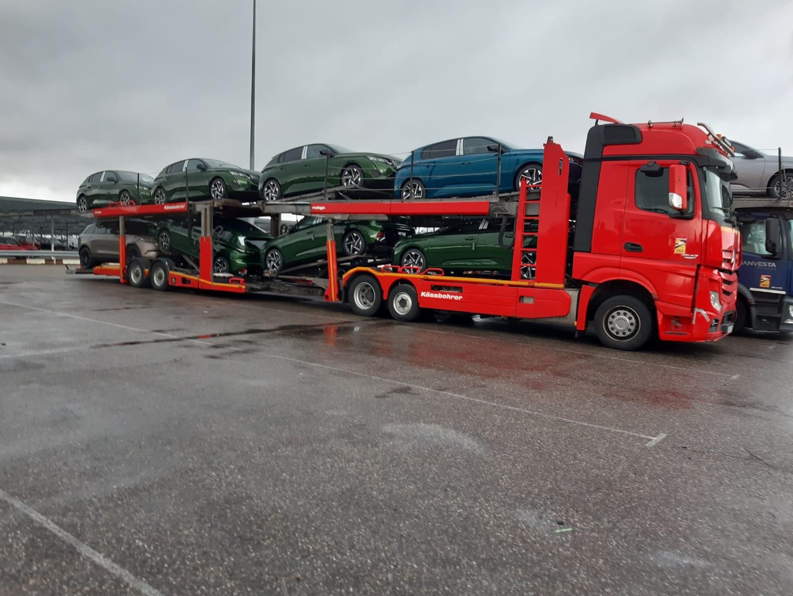 Czerwona laweta Kässbohrer z zielonymi i niebieskimi samochodami osobowymi na mokrym placu, widok z boku, pochmurny dzień.