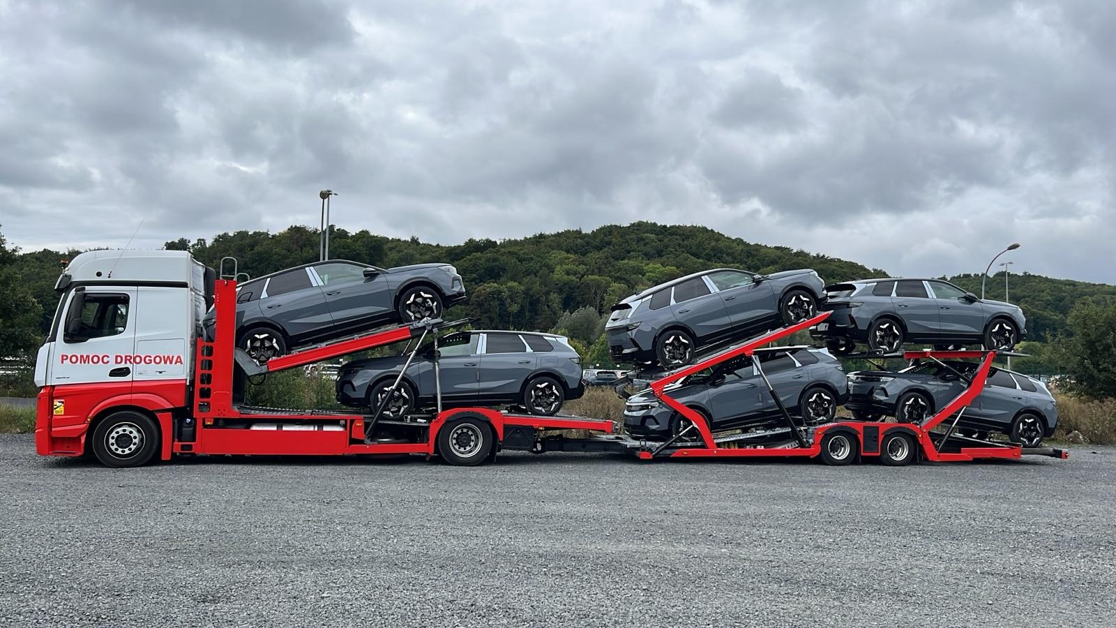 Czerwono-biała laweta z napisem 'Pomoc Drogowa' przewozi sześć szarych samochodów osobowych na tle pochmurnego nieba i zieleni wzgórz.