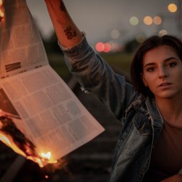 TMK PHOTOVIDEO - Kobieta w dżinsowej kurtce trzymająca płonący egzemplarz gazety na tle torów kolejowych i rozmytych świateł miasta o zmierzchu. Widoczne tatuaże na ramieniu.