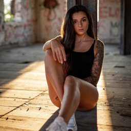 TMK PHOTOVIDEO - Portret kobiety z tatuażem na ramieniu, siedzącej na podłodze w opuszczonym budynku, oświetlonej promieniami słońca. Ubrana w czarny strój i białe buty. Graffiti w tle.