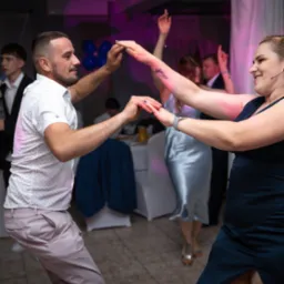 Uśmiechnięci ludzie tańczą na imprezie, trzymając się za ręce. W tle widoczny stół z napojami i dekoracje. Oświetlenie sceniczne tworzy radosną atmosferę.