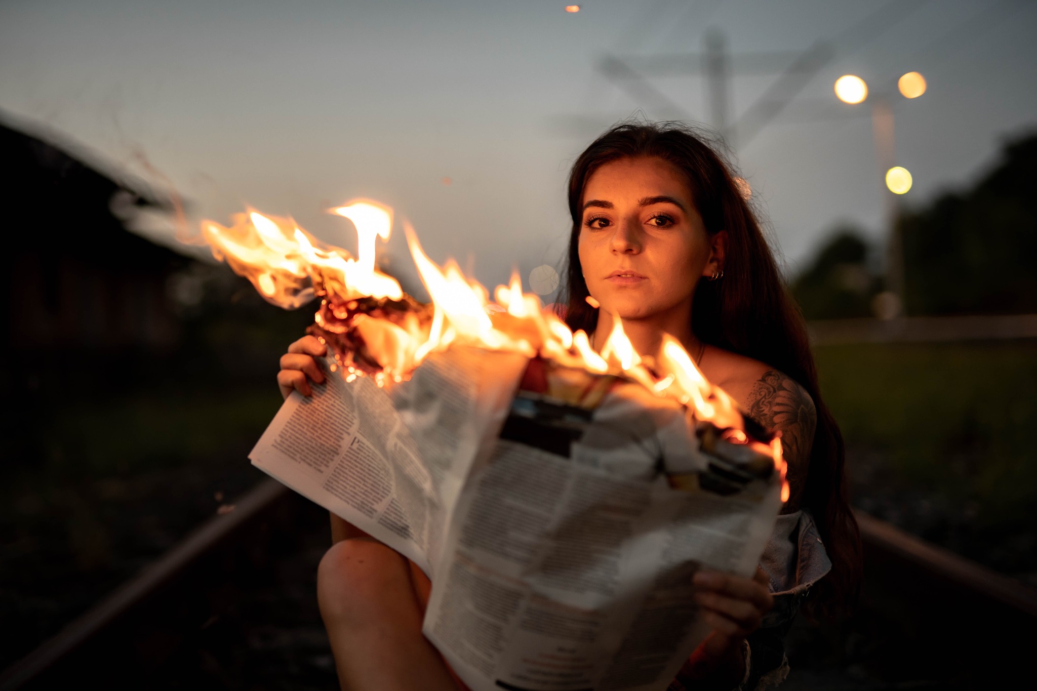 Kobieta z tatuażem na ramieniu trzyma płonący egzemplarz gazety na tle torów kolejowych o zmierzchu, z widocznymi światłami w oddali.