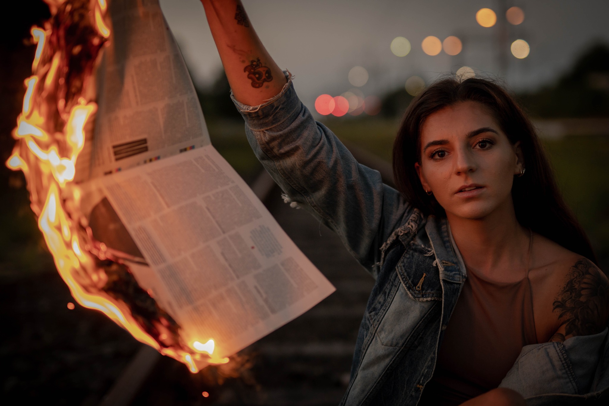 Kobieta w dżinsowej kurtce trzymająca płonący egzemplarz gazety na tle torów kolejowych i rozmytych świateł miasta o zmierzchu. Widoczne tatuaże na ramieniu.