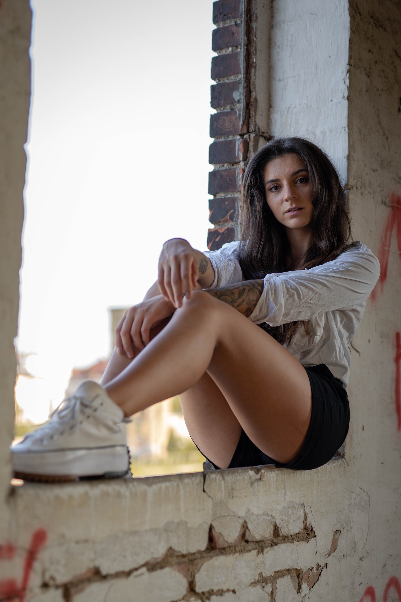 Portret kobiety z tatuażami, siedzącej w oknie z ceglaną ramą. Ubrana w białą koszulę i czarne spodenki, patrzy w obiektyw. Lekkie, naturalne światło.