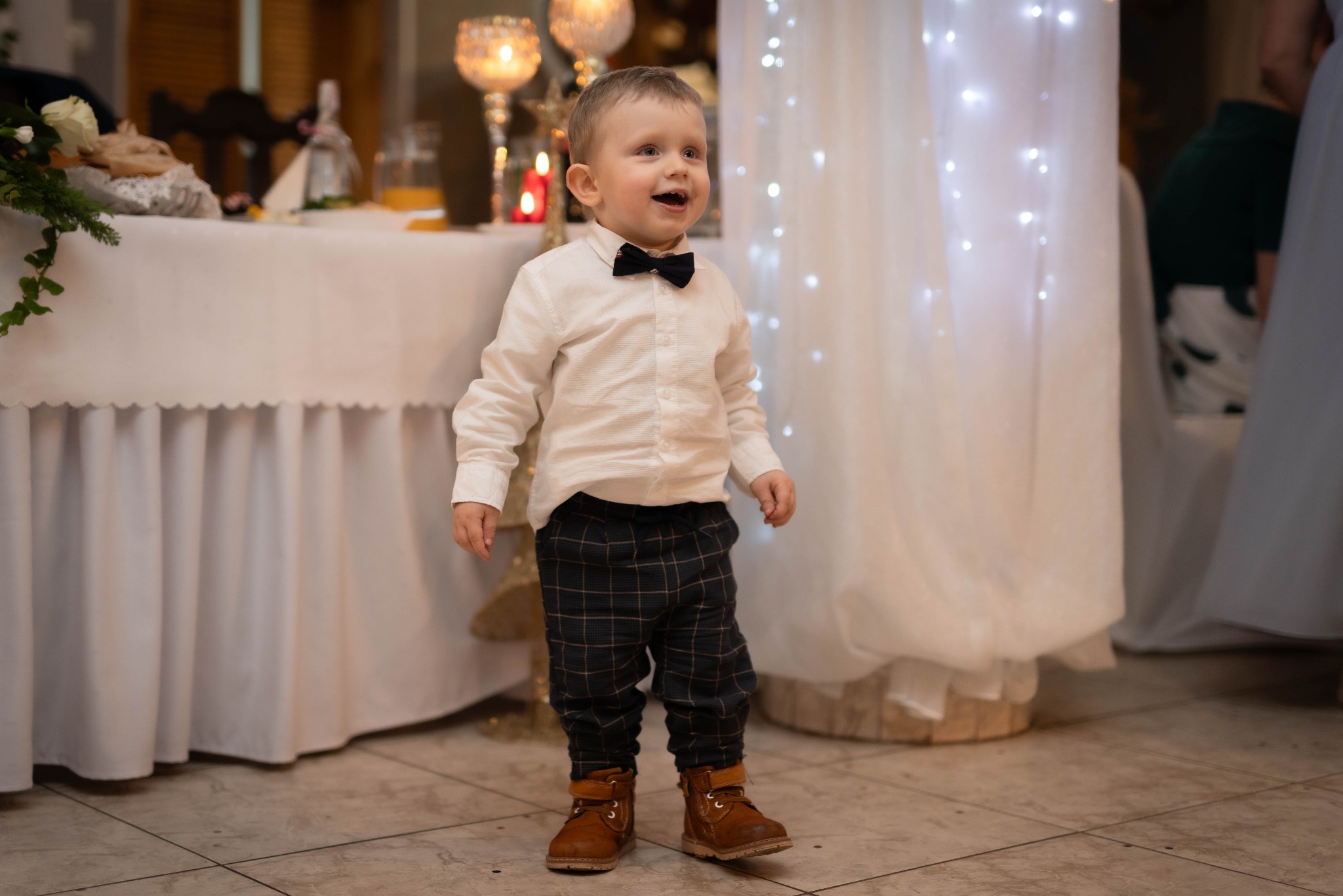 Uśmiechnięty chłopiec w muszce i eleganckim stroju, stojący na tle udekorowanego stołu. Scena z uroczystości, możliwe, że wesela.