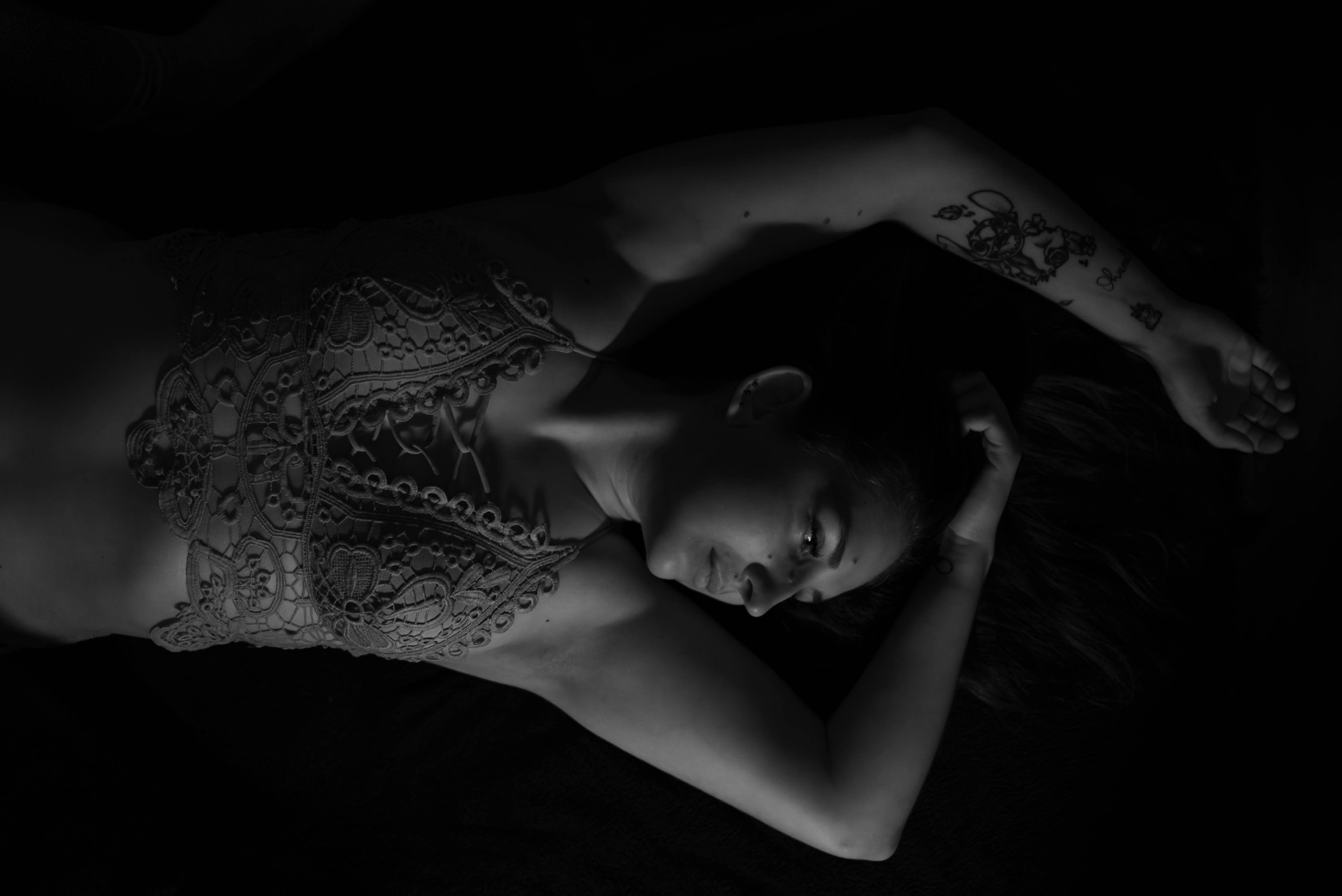 Kobieta w koronkowej bieliźnie leży na ciemnym tle, z tatuażami na ramieniu. Zdjęcie czarno-białe, z delikatnym oświetleniem akcentującym detale.