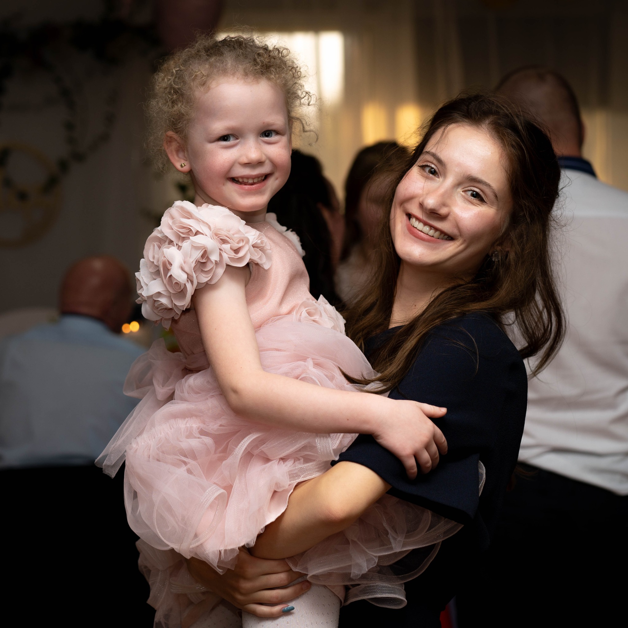 Uśmiechnięta dziewczynka w różowej sukience na rękach kobiety, w tle osoby i dekoracje. Zdjęcie wykonane w ciepłym oświetleniu, z bliska, z widocznymi detalami sukienki.